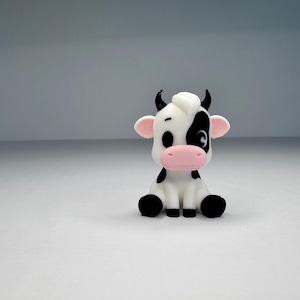 Könnte beinhalten: Eine kleine, sitzende Kuhfigur mit weißem Körper und schwarzen Flecken, einer rosa Schnauze und rosa Innenohren. Die Kuh hat schwarze Hörner, schwarze Augen und eine weiße Haartolle auf dem Kopf in einem cartoonartigen, stilisierten Design.