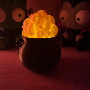 May include: A small black cauldron filled with a cluster of translucent, glowing orange spheres that emit a warm light. The cauldron has a rounded base and two small side handles, positioned between two stylized character figures in the background.