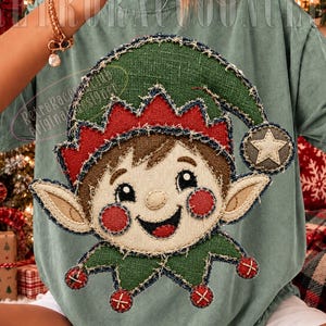 Puede incluir: Una camiseta verde salvia con una gran aplicación texturizada de un elfo sonriente con cabello castaño, mejillas rojas y un sombrero verde con una estrella. El diseño tiene un aspecto deshilachado y cosido. El texto RetroRaccoonClub es visible.