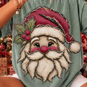 Puede incluir: Una camiseta verde salvia con un gráfico grande y texturizado de Santa Claus con un gorro rojo y una ramita de acebo. El diseño tiene un aspecto deshilachado similar al yute con costuras visibles y una nariz roja prominente.