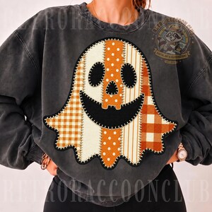 May include: A charcoal gray crewneck sweatshirt featuring a large central graphic of a ghost made of vertical fabric strips in orange and white patterns, including polka dots, stripes, and gingham, with black felt-like eyes and a wide smiling mouth.