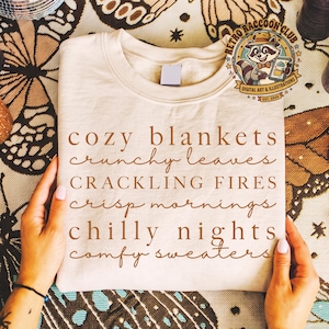 May include: A cream-colored t-shirt featuring brown text that reads: cozy blankets, crunchy leaves, CRACKLING FIRES, crisp mornings, chilly nights, comfy sweaters. A circular logo in the top right corner depicts a raccoon with the text RETRO RACCOON CLUB.