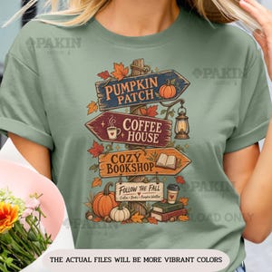 Può includere: Una t-shirt verde salvia con la grafica di un cartello stradale in legno con frecce direzionali. I cartelli riportano le scritte PUMPKIN PATCH, COFFEE HOUSE, COZY BOOKSHOP e FOLLOW THE FALL. Il design include illustrazioni di zucche, una lanterna, una tazza di caffè, un libro aperto e foglie autunnali.