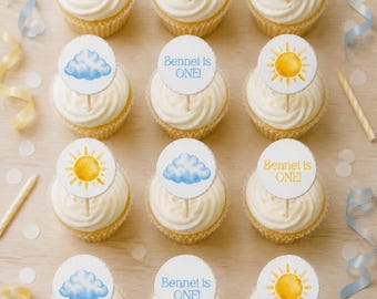 May include: Twelve cupcakes with white frosting, each topped with a circular paper topper featuring watercolor illustrations of yellow suns, blue clouds, or the text Bennet Is ONE! in yellow or blue, arranged in a grid on a light surface.
