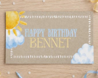 May include: A rectangular birthday card on a wooden surface with light blue and yellow ribbons and candles. The card features a watercolor sun, blue clouds, a white dashed border, and the text "HAPPY BIRTHDAY BENNET" in blue and gold.