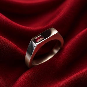Può includere: Un anello in metallo color argento con una superficie superiore piatta e rettangolare, caratterizzato da una singola piccola pietra preziosa rossa di forma rettangolare incastonata orizzontalmente al centro della fascia liscia e lucida.