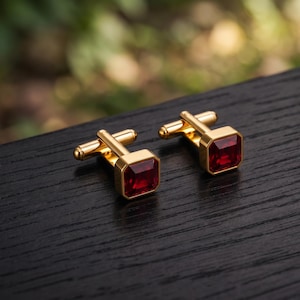May include: A pair of gold-toned cufflinks featuring a square-cut, deep red gemstone set in the center. Each cufflink has a polished gold metal frame and a standard swivel-style toggle mechanism on the back for securing through a shirt cuff.