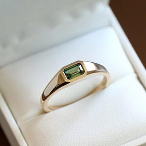 Puede incluir: Un anillo de oro con una piedra preciosa verde de corte esmeralda rectangular engastada en una banda de metal lisa y pulida. El anillo presenta un diseño sólido y elegante con un engaste de bisel que asegura la piedra preciosa.