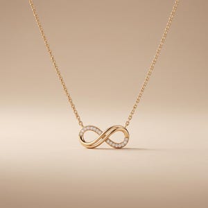 Puede incluir: Un collar de tono dorado con un colgante en forma de símbolo de infinito. El colgante está formado por dos bucles entrelazados, uno de los cuales está adornado con una serie de pequeñas piedras redondas y transparentes en el borde exterior.