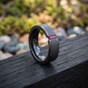 May include: A dark grey matte finish ring featuring a single rectangular red gemstone inlay set horizontally into the band. The ring has a smooth, wide profile with bevelled edges and a polished interior surface that reflects the surrounding environment.