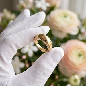 May include: A polished gold band ring with a smooth, rounded surface and a high-shine finish. The ring features a thick, solid construction with a curved profile, showcasing a reflective metallic texture that catches the light evenly.