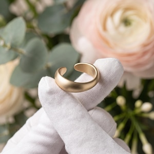 Könnte beinhalten: Ein goldfarbener Ring mit glatter, matter Oberfläche und offenem Design. Das Band hat ein abgerundetes, dickes Profil, das sich zu einer Kreisform biegt und einen kleinen Spalt zwischen den beiden Enden des Metallbandes für eine Passform lässt.