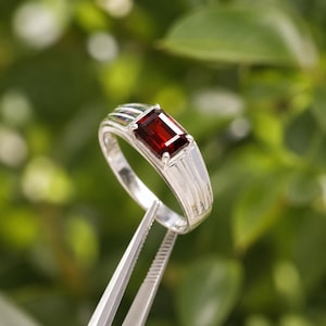May include: A silver ring featuring a rectangular, emerald-cut deep red garnet gemstone set in a four-prong mounting. The band has a polished metallic finish with three distinct vertical grooves on each side of the central stone setting.