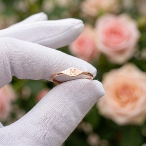 May include: A rose gold ring featuring a small, flat, oval-shaped face engraved with the capital letter M. The band is thin and polished, showcasing a metallic finish that reflects light. The ring is held against a soft, out-of-focus floral background.