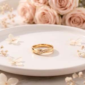 May include: A gold band ring featuring a smooth, polished finish and a diagonal channel set with a row of small, round, clear stones. The ring is centred on a white plate surrounded by soft, light pink roses and small white floral accents.