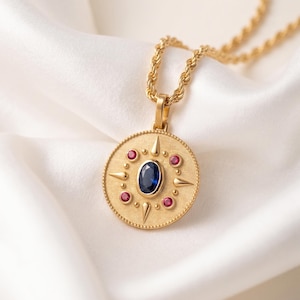 May include: A gold pendant necklace featuring a circular medallion with a central oval blue sapphire, surrounded by four small round red rubies and four pointed gold spikes. The medallion has a beaded edge and hangs from a gold rope chain.
