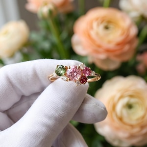 May include: A gold ring featuring a cluster of round-cut gemstones in shades of pink, red, and green, accented by small clear stones. The central arrangement includes a large pink stone, a green stone, and a deep red stone, set on a thin band.