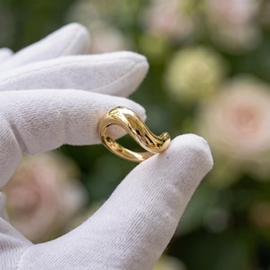 May include: A polished gold ring featuring a smooth, continuous wave-like design that curves upward. The metal surface is highly reflective, showcasing a bright yellow-gold tone with fluid, rounded edges that create an open, asymmetrical silhouette.