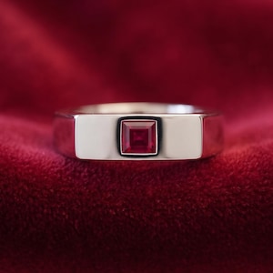 May include: A polished silver-toned metal ring featuring a single square-cut deep red gemstone set flush into the centre of the band. The band has a smooth, wide, and flat surface design that highlights the vibrant red square stone.