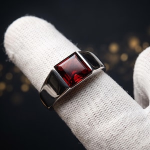 Könnte beinhalten: Ein silberfarbener Metallring mit einem großen, quadratisch geschliffenen, tiefroten Edelstein in der Mitte. Die Ringschiene ist glatt und poliert und zeigt ein klares, massives Design, das die facettierte Oberfläche des dunkelroten Steins betont.