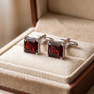 Può includere: Un paio di gemelli quadrati con granato rosso intenso incastonati in montature di metallo color argento lucido. Ogni gemello presenta una pietra preziosa sfaccettata con un'incastonatura a quattro griffe e un meccanismo a perno sul retro.