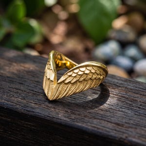 Puede incluir: Un anillo de tono dorado con un diseño detallado de dos alas simétricas que se encuentran en un punto central. La superficie metálica muestra intrincadas texturas de plumas grabadas en toda la extensión de las alas, creando una silueta en V.