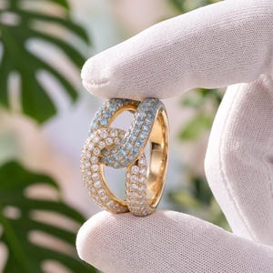 Puede incluir: Un anillo de oro con un diseño de eslabones entrelazados. La superficie está engastada con numerosas piedras pequeñas de corte redondo, con una sección de piedras azul claro y otra sección de piedras blancas transparentes en el aro.