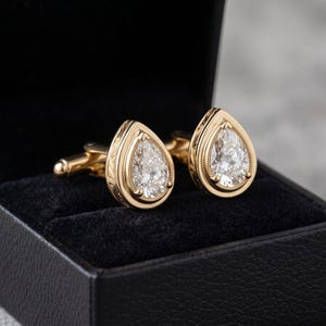May include: A pair of gold cufflinks featuring pear-shaped clear stones set in a bezel with milgrain detailing. The cufflinks have a polished gold finish and are displayed inside a black velvet-lined box, showcasing a teardrop design.