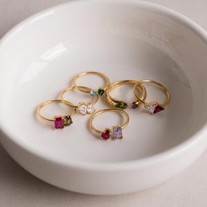 Può includere: Una collezione di anelli color oro con varie pietre preziose colorate in diverse forme, inclusi tagli rettangolari, a cuore e triangolari. Gli anelli sono disposti insieme, mostrando diverse combinazioni di pietre rosa, verdi, blu e trasparenti.