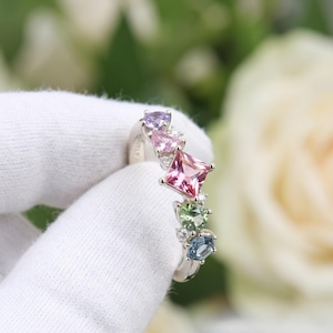 May include: A silver ring featuring a cluster of multi-colored gemstones including a purple stone, two pink stones of different shapes, a green stone, and a blue stone, accented with small clear stones set along a polished silver band.