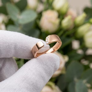 Könnte beinhalten: Ein offener Ring aus Roségold mit einer glatten, polierten Ringschiene und einem kleinen, geschwungenen Akzentbereich, der mit drei winzigen, klaren Rundschliff-Steinen besetzt ist. Der Ring hat eine Lücke zwischen den Enden.