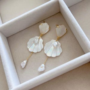 Könnte beinhalten: Ein Paar Ohrhänger mit goldenem Stecker und einem weißen, irisierenden Anhänger in Muschelform. In der Mitte jeder Muschel befindet sich eine kleine, unregelmäßige weiße Perle, an einer feinen Goldkette hängt jeweils eine weitere Perle.