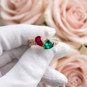 May include: A gold ring featuring a pear-cut red ruby and a pear-cut green emerald set side-by-side. The band is split into two thin, parallel rows, each encrusted with small, clear, round-cut white diamonds that extend along the shank.