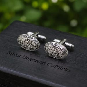 Puede incluir: Un par de gemelos ovalados de plata con un intrincado patrón floral y de volutas grabado. Los gemelos se muestran sobre una caja de madera oscura con el texto Silver Engraved Cufflinks grabado en la superficie con una fuente serif.