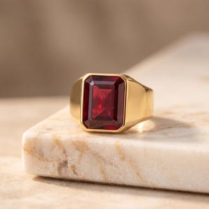 Puede incluir: Un anillo de sello de oro con una gran piedra preciosa de color rojo intenso, rectangular y facetada en el centro. La banda es lisa y pulida, con una construcción gruesa y sólida que se estrecha ligeramente hacia la parte posterior.