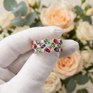 Pode incluir: Um anel de banda dourada com uma disposição de várias filas de pedras preciosas de corte marquesa em vermelho, azul e verde, intercaladas com pequenas pedras redondas transparentes. O design cria um padrão denso e colorido na banda.