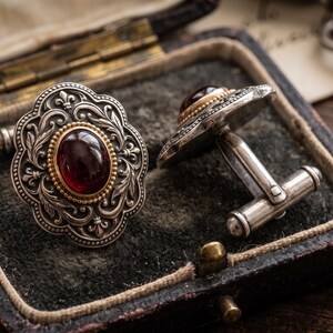 Puede incluir: Un par de gemelos de plata con una piedra preciosa ovalada de color rojo oscuro engastada en un borde de cuentas dorado. La cara de plata tiene un borde festoneado con motivos florales y de flor de lis, unido a un poste en forma de T.