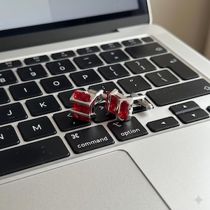 Op de afbeelding: Een paar zilverkleurige manchetknopen met rechthoekige, gefacetteerde rode stenen. De manchetknopen liggen op een zwart laptoptoetsenbord met witte letters, inclusief de toetsen met de tekst command en option.