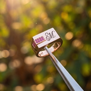 Può includere: Un anello a sigillo in oro con una faccia rettangolare incisa con la lettera M in carattere corsivo. Un piccolo inserto rettangolare sul lato della testa dell'anello è incastonato con sei piccole pietre rosa disposte in due file da tre.
