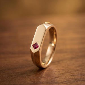 Könnte beinhalten: Ein polierter Goldring mit einem glatten, flachen, rechteckigen Siegeldesign. Ein einzelner, kleiner, quadratisch geschliffener, leuchtend roter Edelstein ist sicher in die Mitte der flachen Oberfläche eingelassen und sorgt für eine klare Optik.