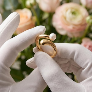 Op de afbeelding: Een goudkleurige open ring met een gladde, gebogen band die eindigt in een enkele, kleine, ronde witte parel. De ring heeft een gepolijst, reflecterend oppervlak en een bypass-ontwerp waarbij de uiteinden elkaar niet raken, wat een opening creëert.