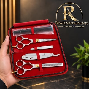 Op de afbeelding: Een rode etui met ritssluiting met een zwarte plastic kam, twee zilveren kappersscharen met gouden accenten, een zilveren scheermes en een zilveren uitdunschaar. De etui bevat de tekst RIAVEENINTRUMENTS QUALITY PRECISION PROFESSIONAL.