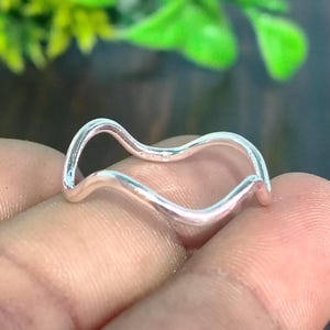 Può includere: Un anello color argento caratterizzato da un design a fascia continua, liscia e ondulata. Il metallo presenta una finitura lucida e riflettente che cattura la luce, creando una forma semplice e fluida che avvolge il dito con un motivo ritmato.