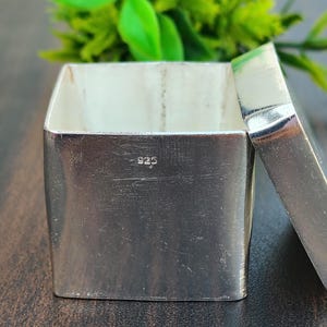 Puede incluir: Una caja cuadrada de plata con tapa extraíble y acabado metálico pulido. El lateral de la caja tiene grabado claramente el sello 925, que indica una composición de plata de ley. El recipiente se encuentra abierto, mostrando su interior hueco.