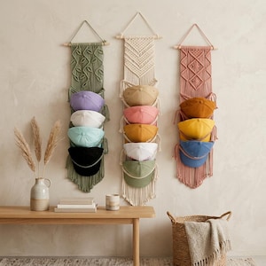 May include: Three vertical macrame wall hangings made of sage green, cream, and dusty rose cord, each featuring wooden dowels and loops designed to hold four baseball caps in various colours including lavender, white, teal, black, tan, pink, yellow, and blue.