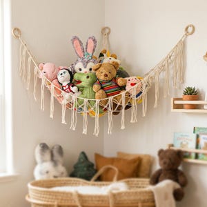 May include: A cream-colored macrame toy hammock suspended in a corner, featuring knotted cord construction with decorative fringe along the bottom edge. The hammock is filled with various plush stuffed animals, including a brown bear.