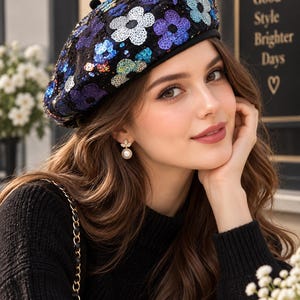 May include: A black beret featuring a dense pattern of embroidered flowers made from iridescent sequins in shades of blue, purple, and silver. The hat has a solid black base fabric and a small black pom-pom at the top center of the crown.