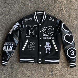 Könnte beinhalten: Eine schwarze Collegejacke mit weiß gestreiftem Rippstrick an Kragen, Ärmelbündchen und Saum. Die Vorderseite ist mit weißem Sticktext und Aufnähern versehen, darunter "Meeeny Club", "MC", "URENEW" und "Creativity is The Substance In Having Melanin."