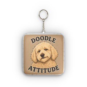 Pode incluir: Um porta-chaves quadrado com uma ilustração de um cão de pelo encaracolado bege sobre um fundo castanho claro. O texto DOODLE ATTITUDE está impresso em letras maiúsculas castanho-escuras acima e abaixo do cão. Inclui um anel de metal prateado.