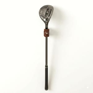May include: A golf hybrid club with a grey metal head and a dark grey shaft, secured to a wall by a brown leather strap mount with a silver metal snap button. The club features a textured grip at the bottom and a detailed metallic club head.